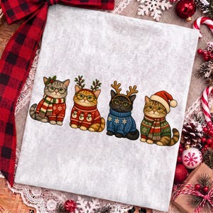 K&ouml;nnte beinhalten: Ein hellgraues T-Shirt mit einer Grafik von vier Katzen, die Brillen und Weihnachtspullover tragen. Die erste Katze tr&auml;gt einen rot-gr&uuml;nen Schal, die zweite ein Rentiergeweih, die dritte ein Geweih und einen blauen Pullover, die vierte eine Weihnachtsm&uuml;tze.