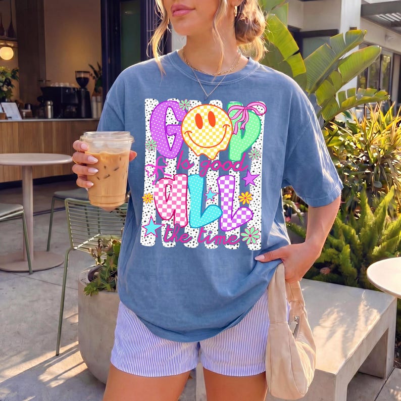Op de afbeelding: Een blauw t-shirt met korte mouwen en een kleurrijke grafische print met de tekst God is good all the time. Het ontwerp bevat een geel geblokt smileygezicht, roze en blauw geblokte letters en kleine bloem- en steraccenten in het patroon.