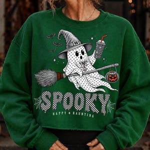 May include: A dark green long-sleeve sweatshirt featuring a graphic of a ghost wearing a witch hat, riding a broomstick, and holding a drink. The ghost has a small pumpkin and bats nearby. The text reads SPOOKY and HAPPY HAUNTING in white.