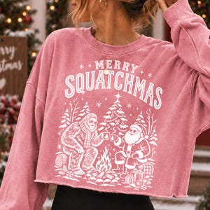 K&ouml;nnte beinhalten: Ein altrosa Langarm-Sweatshirt mit offenem Saum und einem wei&szlig;en Grafikdruck von Bigfoot und dem Weihnachtsmann, die an einem Lagerfeuer sitzen. &Uuml;ber der Illustration steht der Text MERRY SQUATCHMAS in wei&szlig;er Schrift.
