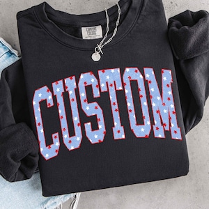 Op de afbeelding: Zwart sweatshirt met het woord "CUSTOM" in grote, lichtblauwe letters gevuld met rode en witte sterren. Een zilveren ketting met een ronde hanger is over het sweatshirt gedrapeerd.