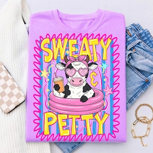 May include: A lilac t-shirt features a cartoon cow in a pink inner tube, wearing heart-shaped sunglasses and a pink bow. The cow holds a drink with a straw. The words "SWEATY PETTY" are in yellow and pink.