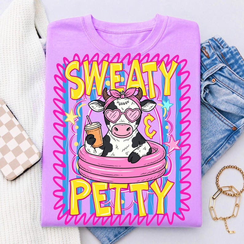 May include: A lilac t-shirt features a cartoon cow in a pink inner tube, wearing heart-shaped sunglasses and a pink bow. The cow holds a drink with a straw. The words "SWEATY PETTY" are in yellow and pink.