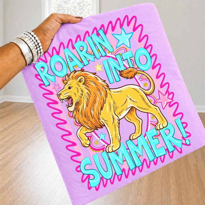 May include: A light purple blanket with a cartoon lion illustration and the words "ROARIN' INTO SUMMER!" in turquoise. The lion is yellow with an orange mane and tail. Pink stars and squiggly lines surround the text and lion.