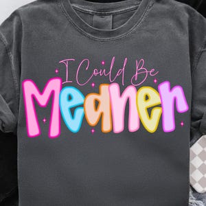 May include: Dark grey sweatshirt featuring the phrase "I Could Be Meaner". The text is in pink script and colourful block letters. The block letters are outlined in pink, with blue, peach, yellow, and purple filling the letters.