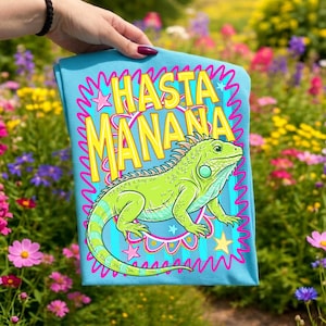 K&ouml;nnte beinhalten: Hellblaues T-Shirt mit einer Illustration einer gr&uuml;nen Leguan-Echse und dem gelben Schriftzug "HASTA MANANA". Die Leguan-Zeichnung ist detailliert, mit einem gestreiften Schwanz. Der Text ist rosa und blau umrandet.