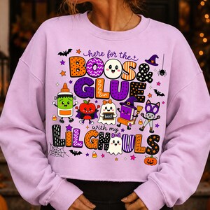 K&ouml;nnte beinhalten: Ein hellviolettes Langarm-Sweatshirt mit einer bunten Halloween-Grafik und dem Text "here for the BOOS & GLUE with my LIL GHOULS". Das Design zeigt Zeichentrickfiguren wie eine Klebeflasche, ein Gespenst und eine Katze sowie Flederm&auml;use und Sterne.
