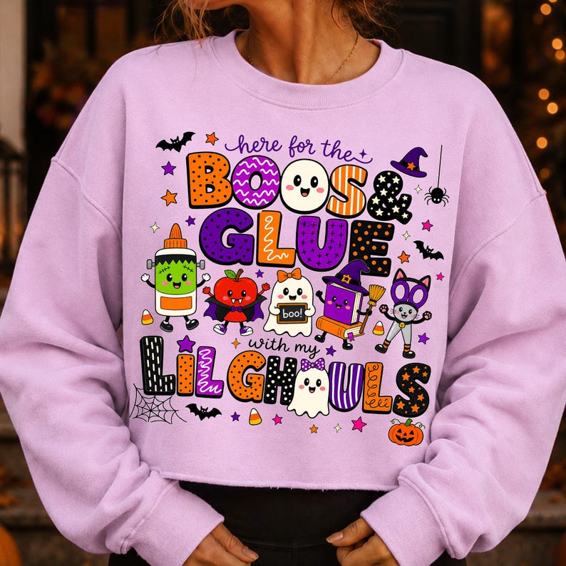 K&ouml;nnte beinhalten: Ein hellviolettes Langarm-Sweatshirt mit einer bunten Halloween-Grafik und dem Text "here for the BOOS & GLUE with my LIL GHOULS". Das Design zeigt Zeichentrickfiguren wie eine Klebeflasche, ein Gespenst und eine Katze sowie Flederm&auml;use und Sterne.