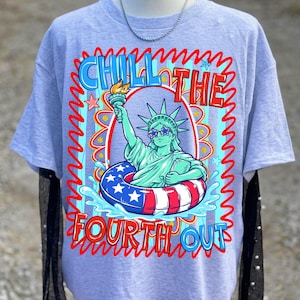 May include: A light gray t-shirt featuring a graphic of the Statue of Liberty wearing star-shaped sunglasses and floating in a red, white, and blue inner tube. The text "CHILL THE FOURTH OUT" is displayed in a wavy font.