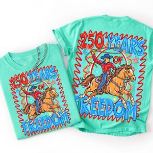 Puede incluir: Dos camisetas turquesas con un dise&ntilde;o gr&aacute;fico. El dise&ntilde;o presenta a un vaquero montando a caballo con un lazo, rodeado por el texto "250 YEARS OF FREEDOM" en rojo y azul. Una camiseta est&aacute; doblada, la otra est&aacute; plana.
