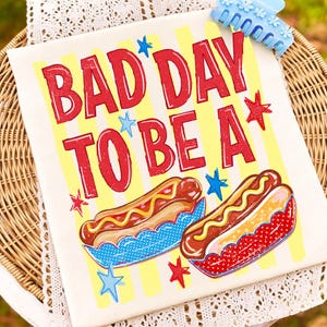 K&ouml;nnte beinhalten: Ein cremefarbenes rechteckiges Zierkissen mit dem Text "BAD DAY TO BE A" in Rot, mit Sternakzenten. Zwei Hotdogs in blauen und roten Schalen sind unten abgebildet. Eine blaue Haarspange befindet sich in der oberen rechten Ecke.