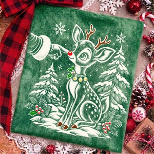 Puede incluir: Un coj&iacute;n cuadrado de terciopelo verde con una ilustraci&oacute;n blanca de un reno de nariz roja brillante siendo tocado por un dedo enguantado. El dise&ntilde;o incluye pinos nevados, copos de nieve, bayas de acebo y un collar con peque&ntilde;as campanas.