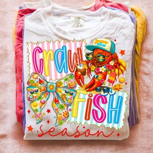 May include: White t-shirt with a colourful graphic design featuring a lobster wearing sunglasses and a hat, with the words "Crawfish Season" in bright colours. A bow with sunglasses is also featured.