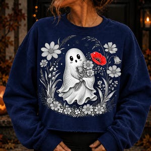 K&ouml;nnte beinhalten: Ein dunkelblaues Langarm-Sweatshirt mit einer zentralen Grafik eines wei&szlig;en Geistes, der einen Blumenstrau&szlig; h&auml;lt. Der Geist ist von einer kreisf&ouml;rmigen Anordnung aus schwarz-wei&szlig;en Strichzeichnungen von Blumen umgeben, inklusive einer roten Mohnblume.