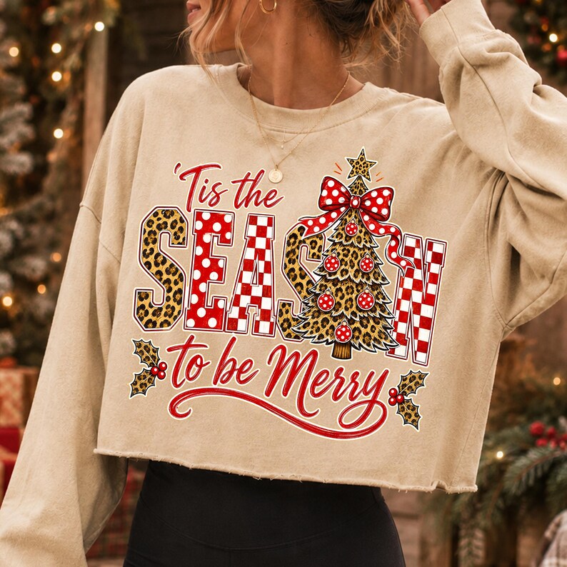 Op de afbeelding: Een beige cropped sweatshirt met lange mouwen en een grafische print met de tekst 'Tis the SEASON to be Merry'. Het ontwerp bevat een luipaardprint, rode en witte stippen, een gestileerde kerstboom en bladeren van hulst.
