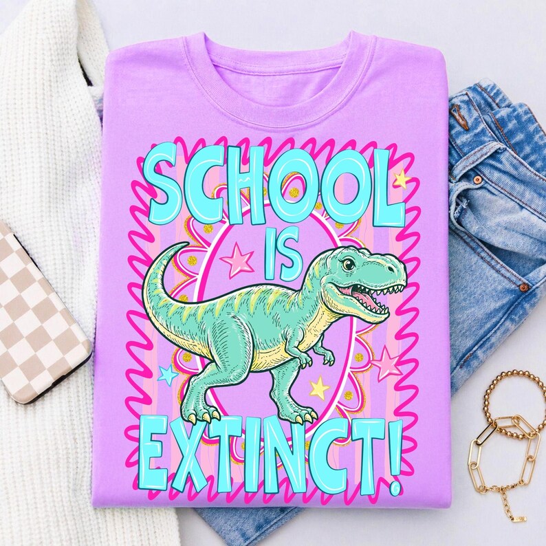 Op de afbeelding: Een lila T-shirt met een afbeelding van een groene Tyrannosaurus Rex en de turquoise tekst "SCHOOL IS EXTINCT!". Het ontwerp bevat roze en gele sterren en een roze rand.