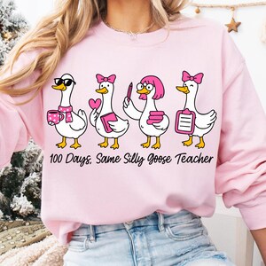 K&ouml;nnte beinhalten: Hellrosa Sweatshirt mit einem Aufdruck von vier Cartoon-G&auml;nsen, jede mit einem einzigartigen Accessoire, wie Sonnenbrille und rosa Schleife. Der Text "100 Days, Same Silly Goose Teacher" ist unter den G&auml;nsen aufgedruckt.