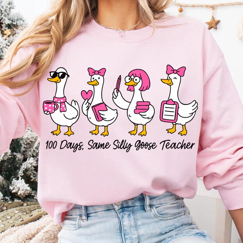 K&ouml;nnte beinhalten: Hellrosa Sweatshirt mit einem Aufdruck von vier Cartoon-G&auml;nsen, jede mit einem einzigartigen Accessoire, wie Sonnenbrille und rosa Schleife. Der Text "100 Days, Same Silly Goose Teacher" ist unter den G&auml;nsen aufgedruckt.
