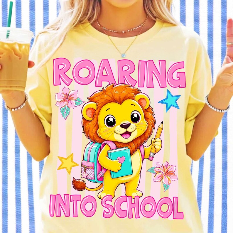 May include: A pale yellow t-shirt featuring a graphic of a cartoon lion holding a blue book and a yellow pencil. The lion wears a pink backpack. The text ROARING INTO SCHOOL is printed in pink. The design includes pink flowers and blue and yellow stars.