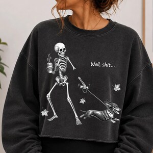 May include: A charcoal gray crewneck sweatshirt featuring a graphic of a skeleton holding a coffee cup and walking a dachshund on a leash. The text "Well, shit..." is printed above the dog, accompanied by small white leaf illustrations.