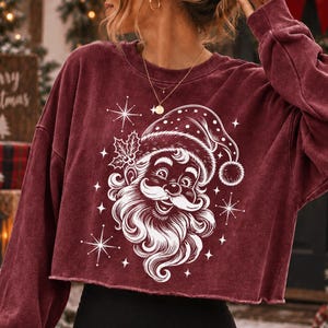 May include: A maroon long-sleeve cropped sweatshirt featuring a white graphic print of a smiling Santa Claus face with a hat, holly leaves, and starburst accents. The garment has a crew neckline and a raw, unfinished hemline at the waist.