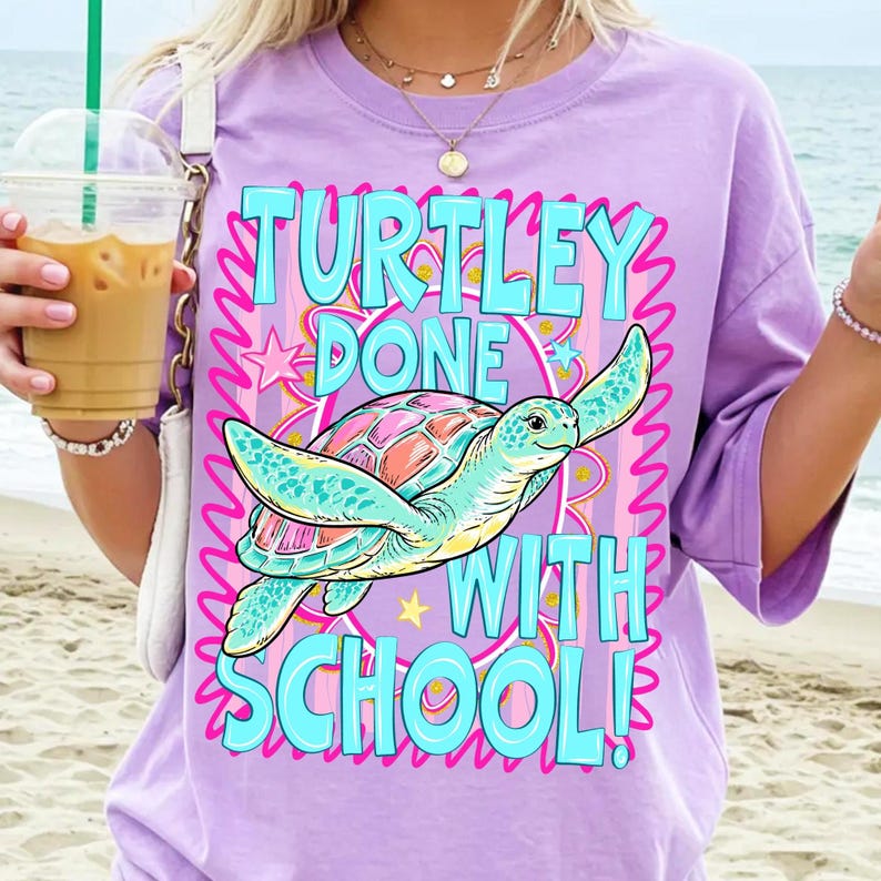 以下が含まれることがあります： 薄紫色のTシャツ。海亀のグラフィックと「TURTLEY DONE WITH SCHOOL！」というターコイズ色の文字がプリントされています。カメはピンクと青色の甲羅とターコイズ色のヒレを持っています。デザインはピンクの波線と星で囲まれています。