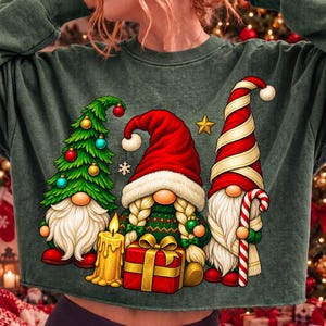 Puede incluir: Una sudadera de manga larga de color verde oscuro con un estampado gr&aacute;fico de tres gnomos. Un gnomo tiene forma de &aacute;rbol de Navidad, otro sostiene una vela amarilla y un regalo rojo, y el tercero lleva un gorro a rayas rojas y blancas con un bast&oacute;n.
