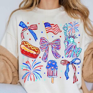 K&ouml;nnte beinhalten: Cremefarbenes Sweatshirt mit patriotischen Illustrationen. Designs umfassen eine amerikanische Flagge, die Freiheitsstatue, Feuerwerk, Schleifen, einen Hotdog und ein Eis am Stiel, alles in Rot, Wei&szlig; und Blau.