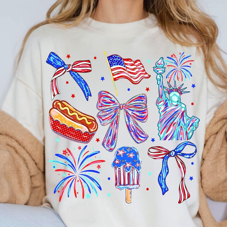 K&ouml;nnte beinhalten: Cremefarbenes Sweatshirt mit patriotischen Illustrationen. Designs umfassen eine amerikanische Flagge, die Freiheitsstatue, Feuerwerk, Schleifen, einen Hotdog und ein Eis am Stiel, alles in Rot, Wei&szlig; und Blau.