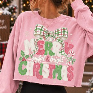K&ouml;nnte beinhalten: Ein hellrosa lang&auml;rmeliges bauchfreies Sweatshirt mit einem gro&szlig;en Grafikdruck einer gr&uuml;n-wei&szlig; karierten Schleife mit einer silbernen Glocke. Der Text lautet Merry joyful & bright Christmas in Rot, Gr&uuml;n und Wei&szlig; mit Stechpalmen.