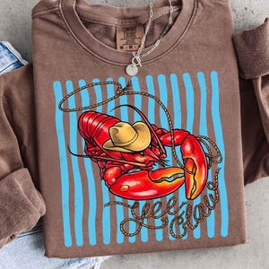Op de afbeelding: Bruin sweatshirt met een afbeelding van een rode kreeft met een gele cowboyhoed, met de tekst "Yee Claw" in een touwachtig lettertype. De kreeft staat tegen een achtergrond van blauwe verticale strepen.