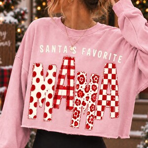 K&ouml;nnte beinhalten: Ein hellrosa lang&auml;rmeliges bauchfreies Sweatshirt mit dem wei&szlig;en Text SANTA'S FAVORITE &uuml;ber dem Wort MAMA. Die Buchstaben in MAMA sind mit verschiedenen roten und wei&szlig;en Mustern gef&uuml;llt, darunter Punkte, Karos, Blumenmuster und Schachbrettmuster.