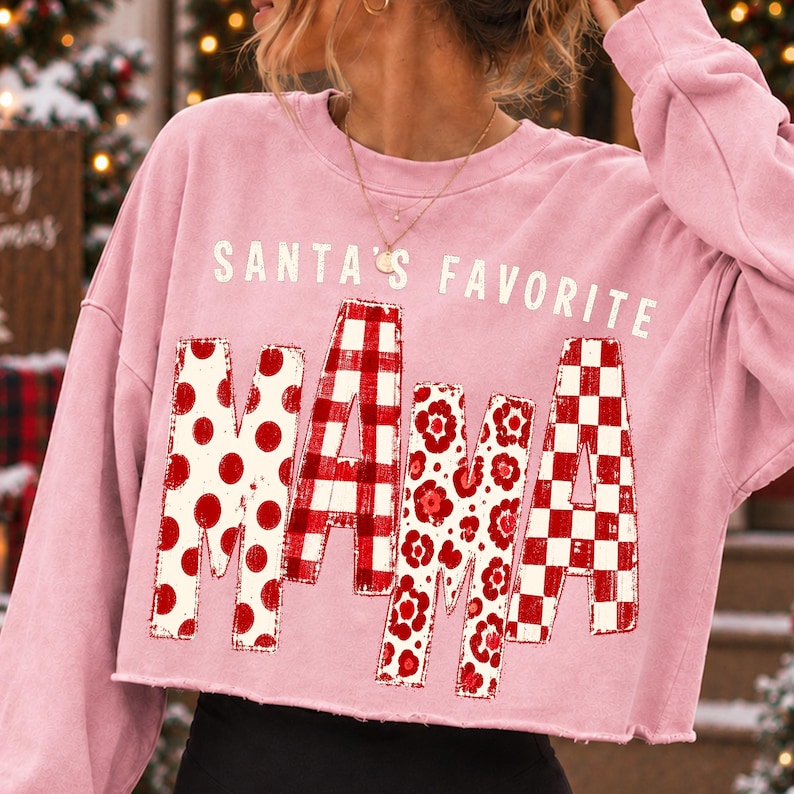 K&ouml;nnte beinhalten: Ein hellrosa lang&auml;rmeliges bauchfreies Sweatshirt mit dem wei&szlig;en Text SANTA'S FAVORITE &uuml;ber dem Wort MAMA. Die Buchstaben in MAMA sind mit verschiedenen roten und wei&szlig;en Mustern gef&uuml;llt, darunter Punkte, Karos, Blumenmuster und Schachbrettmuster.
