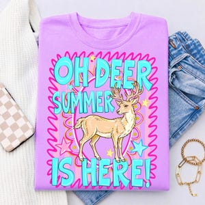 K&ouml;nnte beinhalten: Ein helllila T-Shirt mit dem Text "OH DEER SUMMER IS HERE!" in T&uuml;rkis. Ein Cartoon-Hirsch ist auf dem T-Shirt zentriert. Rosa Sterne und wellenf&ouml;rmige Linien umgeben den Text und den Hirsch. Das T-Shirt ist gefaltet.