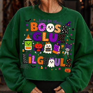 K&ouml;nnte beinhalten: Ein dunkelgr&uuml;nes Langarm-Sweatshirt mit einer bunten Halloween-Grafik und dem Text "here for the BOOS & GLUE with my LIL GHOULS". Das Design zeigt Zeichentrick-Geister, eine Klebeflasche, einen Apfel, ein Buch und eine Katze.