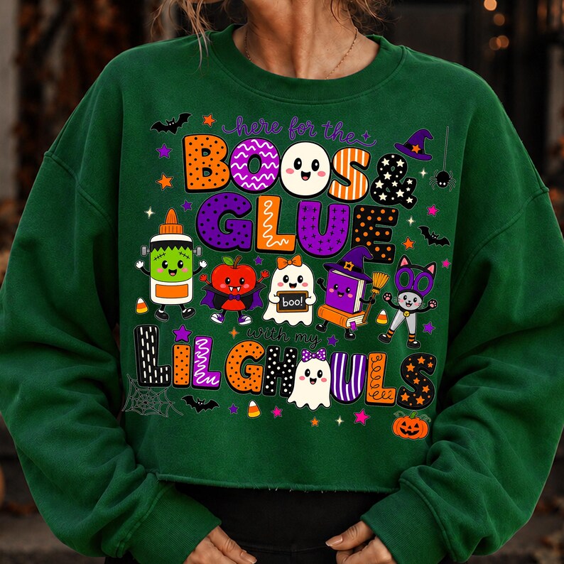 K&ouml;nnte beinhalten: Ein dunkelgr&uuml;nes Langarm-Sweatshirt mit einer bunten Halloween-Grafik und dem Text "here for the BOOS & GLUE with my LIL GHOULS". Das Design zeigt Zeichentrick-Geister, eine Klebeflasche, einen Apfel, ein Buch und eine Katze.