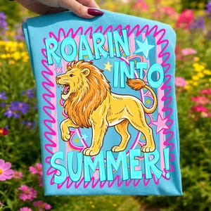May include: A light blue t-shirt featuring a cartoon lion with a golden mane and the text "ROARIN' INTO SUMMER!" in turquoise. The design includes pink accents, stars, and a wavy border. The shirt is held up against a background of colorful flowers.