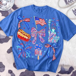 K&ouml;nnte beinhalten: Blaues T-Shirt mit patriotischen Illustrationen. Das Design umfasst eine amerikanische Flagge, die Freiheitsstatue, Feuerwerk, einen Hotdog und ein Eis am Stiel. Das T-Shirt ist mit roten, wei&szlig;en und blauen Akzenten verziert.