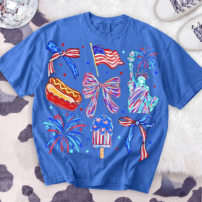 K&ouml;nnte beinhalten: Blaues T-Shirt mit patriotischen Illustrationen. Das Design umfasst eine amerikanische Flagge, die Freiheitsstatue, Feuerwerk, einen Hotdog und ein Eis am Stiel. Das T-Shirt ist mit roten, wei&szlig;en und blauen Akzenten verziert.