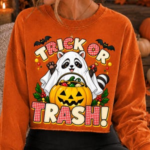 May include: An orange long-sleeve sweatshirt featuring a graphic of a raccoon wearing a white ghost costume. The raccoon holds a jack-o'-lantern bucket filled with sweets. The text reads TRICK OR TRASH! with black bats and green leaves.