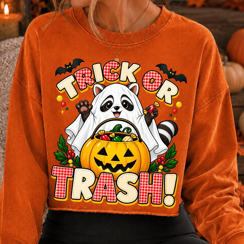 May include: An orange long-sleeve sweatshirt featuring a graphic of a raccoon wearing a white ghost costume. The raccoon holds a jack-o'-lantern bucket filled with sweets. The text reads TRICK OR TRASH! with black bats and green leaves.