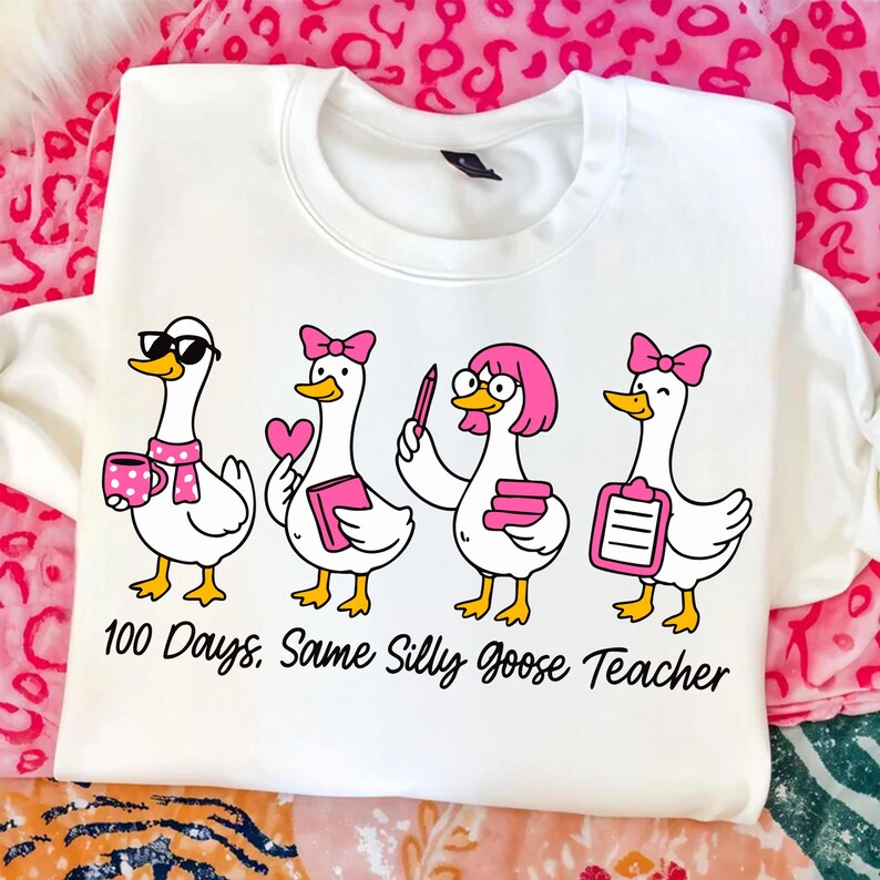 K&ouml;nnte beinhalten: Wei&szlig;es Sweatshirt mit einer Cartoon-Illustration von vier G&auml;nsen, jede mit unterschiedlichen Accessoires wie Sonnenbrillen, Schleifen und B&uuml;chern. Der Text "100 Days. Same Silly Goose Teacher" ist darunter aufgedruckt. Das Design verwendet Schwarz, Rosa und Gelb.