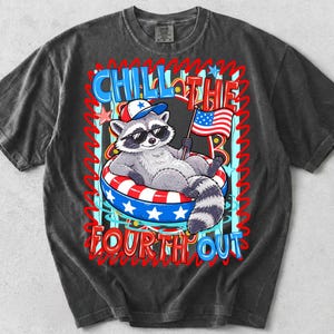 Op de afbeelding: Donkergrijze t-shirt met een cartoon wasbeer met een zonnebril en een patriottische hoed, liggend in een rood, wit en blauwe zwemband. Het shirt heeft de tekst "CHILL THE FOURTH OUT" in een kleurrijk ontwerp.