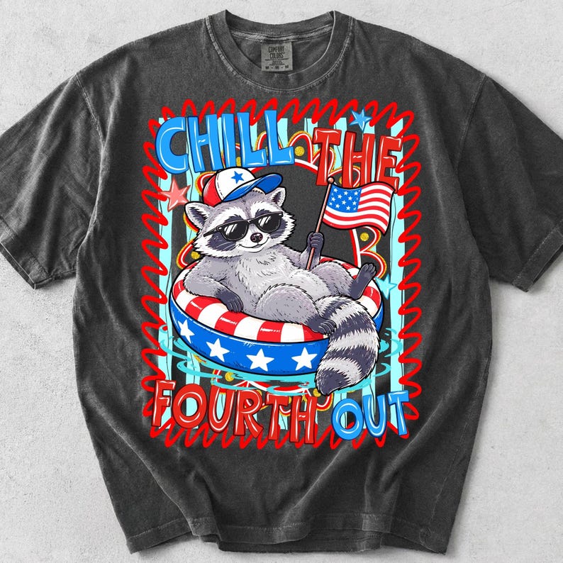 Op de afbeelding: Donkergrijze t-shirt met een cartoon wasbeer met een zonnebril en een patriottische hoed, liggend in een rood, wit en blauwe zwemband. Het shirt heeft de tekst "CHILL THE FOURTH OUT" in een kleurrijk ontwerp.