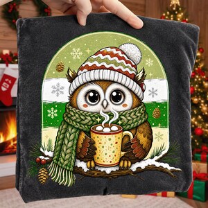 May include: A dark gray t-shirt featuring a graphic of a brown owl wearing a red, white, and green striped knit hat and a green cable-knit scarf. The owl holds a yellow mug with white marshmallows, set against a green and white background.