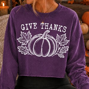 Puede incluir: Una sudadera morada de manga larga con cuello redondo y dobladillo sin rematar. El frente presenta un gr&aacute;fico blanco de una calabaza flanqueada por hojas de arce y roble, con el texto "GIVE THANKS" impreso en una fuente serif blanca arriba.