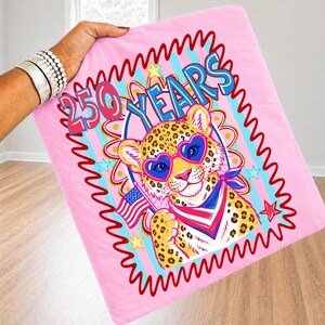 Op de afbeelding: Roze deken met een luipaard met een hartvormige zonnebril en een patriottische bandana, met een Amerikaanse vlag. De deken heeft de tekst "250 YEARS" in een kleurrijk ontwerp, omgeven door een rode golvende rand.