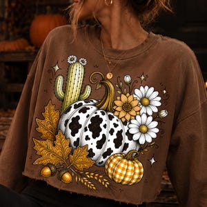 Puede incluir: Una sudadera marr&oacute;n de manga larga con un estampado gr&aacute;fico de una calabaza con manchas de vaca, una calabaza m&aacute;s peque&ntilde;a de cuadros amarillos, un cactus verde con flores blancas, hojas de oto&ntilde;o, bellotas, espigas de trigo y margaritas.
