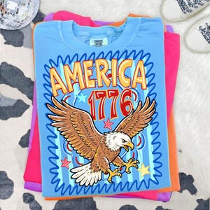 May include: A light blue t-shirt with the word "AMERICA" and the number "1776" in gold and red. The shirt features a brown and white eagle with outstretched wings, surrounded by stars and blue wavy lines. The shirt is folded on top of other colorful shirts.
