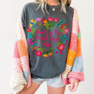 K&ouml;nnte beinhalten: Dunkelgraues Sweatshirt mit floralem Herzdesign und dem Text "Don't Let The Hard Days Win". Das Sweatshirt wird mit einer bunten Patchwork-Strickjacke kombiniert. Das florale Design umfasst rosa, gelbe und blaue Blumen.